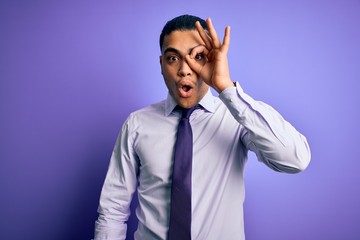 Young brazilian businessman wearing elegant tie standing over isolated purple background doing ok gesture shocked with surprised face, eye looking through fingers. Unbelieving expression.