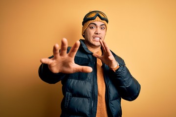 Young brazilian skier man wearing snow sportswear and ski goggles over yellow background afraid and terrified with fear expression stop gesture with hands, shouting in shock. Panic concept.