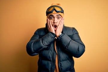 Young brazilian skier man wearing snow sportswear and ski goggles over yellow background afraid and shocked, surprise and amazed expression with hands on face