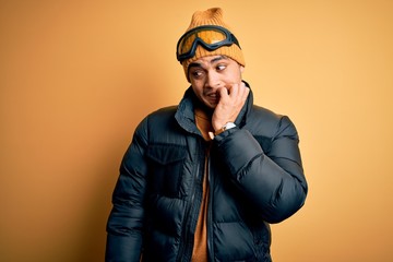 Young brazilian skier man wearing snow sportswear and ski goggles over yellow background looking stressed and nervous with hands on mouth biting nails. Anxiety problem.