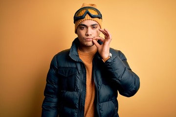 Young brazilian skier man wearing snow sportswear and ski goggles over yellow background mouth and...