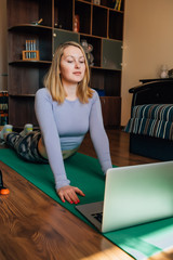 Young woman in yoga pose watching online class in room during quarantine, coronavirus pandemic. Attractive slim girl in sportswear doing fitness exercises on mat. Sport at home, lockdown.