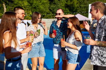 Group of friends having fun at poolside summer party clinking glasses with summer cocktails on sunny day near swimming pool. People toast drinking fresh juice at luxury villa on tropical vacation.