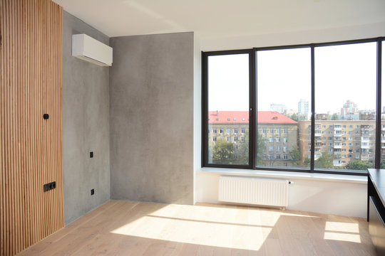 An empty apartment room with a large window and radiator heater, wood paneled and gray painted wall with air conditioner and wood laminated floor.