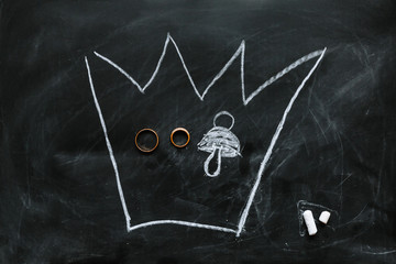 Drawing with chalk on a blackboard. A crown containing wedding rings and a baby nipple