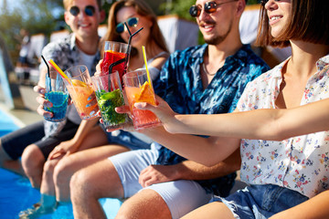 Close view. Friends clinking glasses with fresh colorful cocktails sitting by swimming pool on...