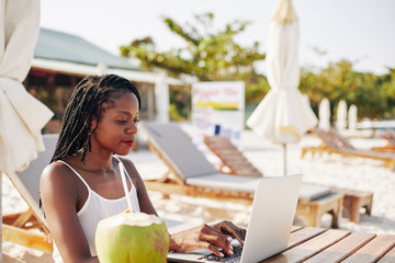 Concentrated female blogger sitting at table on beach and working on article or blog post