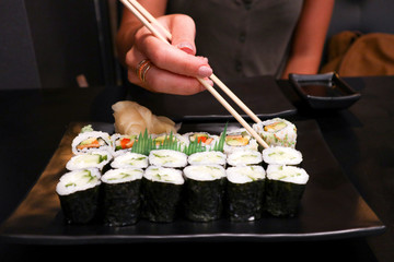 a young girl is sitting in Japanese cafe and eating tasty sushi with chopsticks on a black plate with saucer on the black table