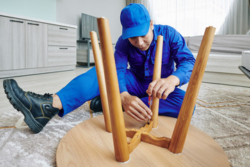 Handyman using screwdriver when assembling wooden table in apartment of customer - obrazy, fototapety, plakaty