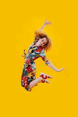 Happy exhilarated woman in a colorful dress jumping in the air over yellow. Arms bent, arms...