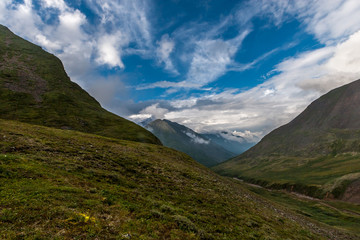 Green hillside and valley between the mountains. Blue sky and low white clouds over the mountain peaks. Horizontal.
