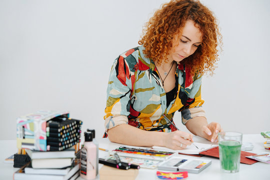 Focused Serious Ginger Artist Woman In Colorful Dress Painting Behind Work Desk In Her Notepad With A Brush. Keeping Her Eyes On A Task.