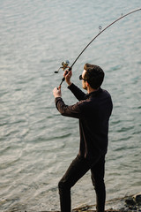Fisherman with rod throws bait into the water on river bank. Fishing for pike, perch, carp. Man catching fish, fishing on lake, pond with text space. Wild nature. The concept of rural getaway.