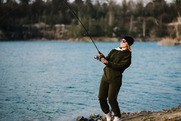 Fisherman with rod, spinning reel on the river bank. Fishing for pike, perch, carp. Woman catching fish, pulling rod while fishing at the weekend. Girl fishing from beach lake or pond with text space.