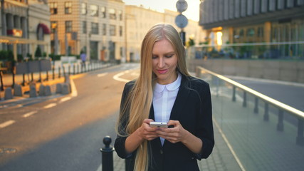 Fototapeta premium Elegant blond woman in suit walking on street and browsing smartphone with smile against urban background.