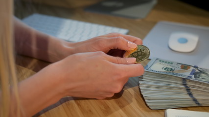 Crop view of hands of woman holding shiny metal bitcoin sitting at wooden desk with wad of cash nearby at night