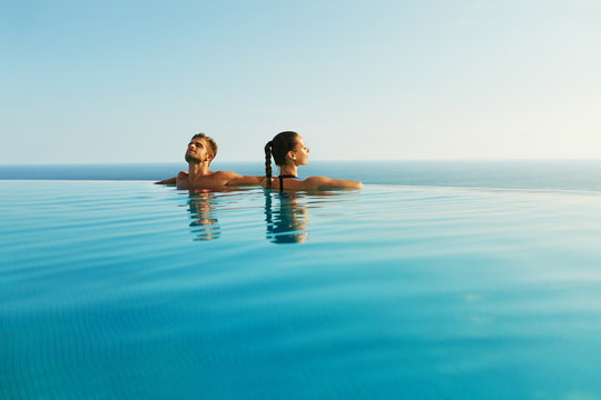 Couple In Love At Luxury Resort On Romantic Summer Vacation. People Relaxing Together In Edge Swimming Pool Water, Enjoying Beautiful Sea View. Happy Lovers On Honeymoon Travel. Relationship, Romance