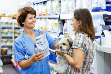 Elderly female veterinarian recommending pet food to young woman visiting pet store with her puppy