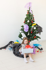 Shocked little child girl looks with opened eyes and worried expression, holding box with various plastic wastes over christmas tree background.
