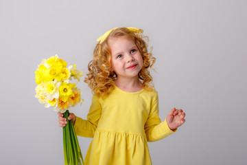 little  curly haired girl in a yellow dress holding a bouquet of spring flowers on a white...