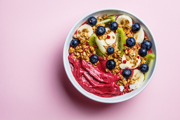 Pink berry smoothie bowl with fruits, berries and granola in a white bowl on pink background, top view. Healthy vegan food concept.