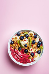 Pink berry smoothie bowl with fruits, berries and granola in a white bowl on pink background, top view