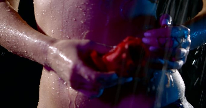 close-up of a female belly in the dark in the rain. the woman runs a red rose over it. drops of water run down the skin. neon light