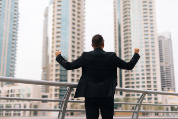 Rear view of young business man in suit with raised hands celebrate win look on skyscrapers