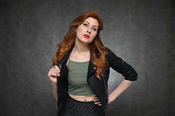 Horizontal portrait of a red-haired young woman in a black jacket and green t-shirt. Model with excellent makeup posing on a gray background in the studio.