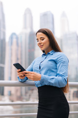 Fototapeta premium Beautiful business woman typing message on a mobile phone while going out the business center in a downtown. Cheerful female looking at a smartphone while standing on a glass building background.
