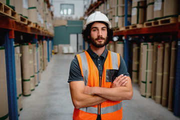 Portrait of storeman in a warehouse for delivering and transporting industrial goods wearing white helmet and orange vest uniform standing with folded arms in aisle - obrazy, fototapety, plakaty