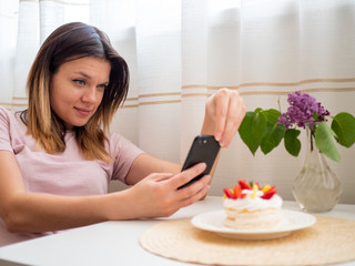 Food blogger woman in pink t-shirt sitting at white table and take photo of cake