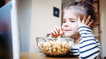 Smiling kid watching cartoons at home using wireless tablet computer and eating pop-corn. Girl toddler growing with gadget in cozy place