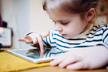 Naklejka premium Pretty little girl playing kid's application using wireless tablet computer. Toddler growing with gadget in cozy place
