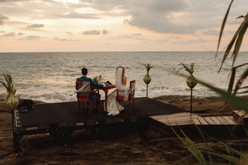 The night wedding ceremony. The bride and groom are sitting at the festive table and looking on sunset by the sea