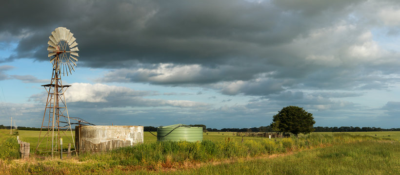 panoramic image showing long stretching green farm fields under a cloudy sky with a wind powered water pump and cement tank in the foreground, Hamilton, rural Victoria, Australia