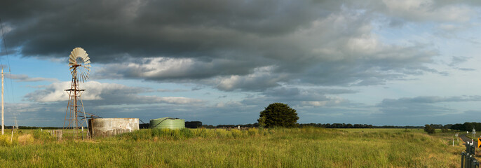 panoramic image showing long stretching green farm fields under a cloudy sky with a wind powered water pump and cement tank in the foreground, Hamilton, rural Victoria, Australia