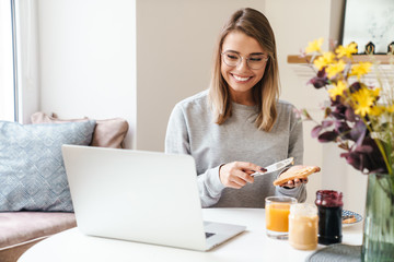 Photo of cheerful woman in eyeglasses eating toasts while using laptop