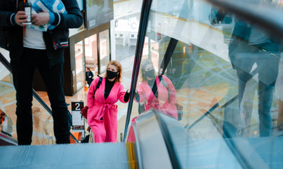 beautiful woman bright coat rides escalator shopping center black protective mask on her face against the virus polluted air.