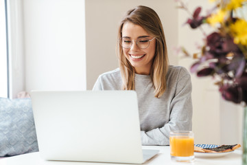 Photo of cheerful young woman using laptop while having breakfast