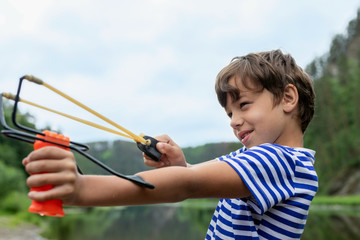 Sly and naughty little boy in striped t-shirt aiming somewhere from slingshot. Close up portrait on...