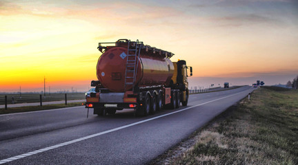 A truck carries a tank of combustible fuel on a highway against a forest and blue sky. The concept of transportation of dangerous goods on the road, license, sunset - obrazy, fototapety, plakaty