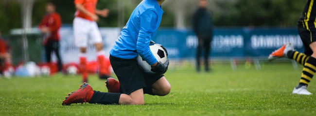 Naklejka premium Young Football Goalkeeper Catching Soccer Ball. Soccer Goalie in Gloves on Knees During Sports Cometition Match