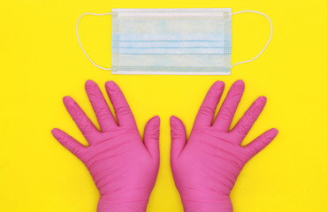 Disposable latex glove. The pink gloves. The nurse's gloved hands are on the table. Disposable surgical blue mask on the table. Security during the quarantine period. Yellow background.