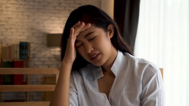 Young beautiful Asian woman having a headache while lying on bed, feeling sad and stressed at home. Disappointed and desperate girl crying.