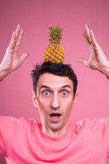 Amazed emotional young man confused with pineapple stand by itself on head. Guy hold hands up. Trick. Isolated over pink background.