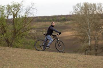 Fototapeta premium Sports brutal bearded guy on a modern mountain bike. Cyclist on the green hills in the spring.