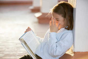 Little girl laughing a lot reading a book sitting on the rooftop
