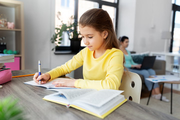 children, education and learning concept - student girl with book writing to notebook and mother working at home