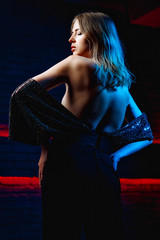 Beautiful girl model in neon lighting. Young woman in a dress with an open back in red with blue lighting in a club
